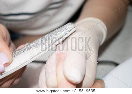 Process manicure close-up. Polishing of nails. Beautician in rubber gloves handles the nail of the client with a nail file. Selective focus on costumer's finger and nail file.