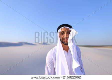 Cheerful Muslim guy walks along white sand of wide desert on warm summer evening. Arab young man in national clothes dances on spot and laughs giggly, adjusting hands with harness in wind. Swarthy, handsome Muslim with short dark hair dressed in kandura, 