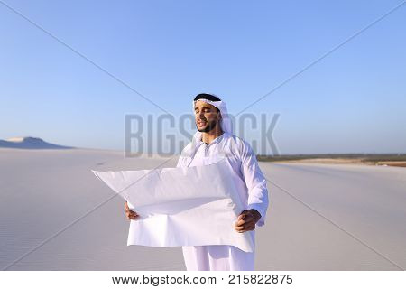 Beautiful Emirate Sheikh businessman studying project of future construction of complex, standing in middle of bottomless desert with white clean sand on clear warm day against blue sky. Swarthy Muslim with short dark hair dressed in kandura, long, spacio