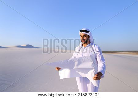 Beautiful Emirate Sheikh businessman studying project of future construction of complex, standing in middle of bottomless desert with white clean sand on clear warm day against blue sky. Swarthy Muslim with short dark hair dressed in kandura, long, spacio