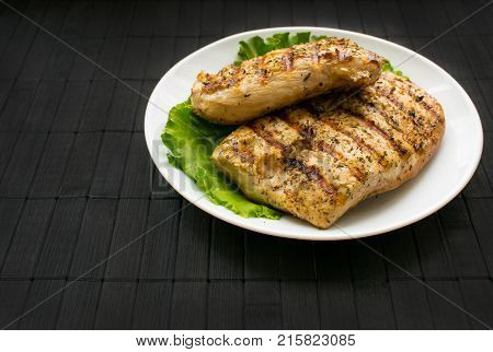 Chicken grilled steak on leaves slate on a white round plate on a black wooden background. Healthy food. Diet, fitness food. B-B-Q.