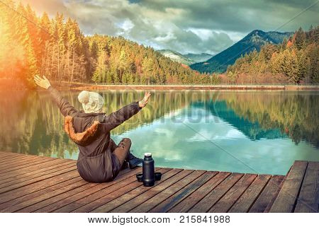 Happy young woman sitting on the wooden pier with hadn up, mountain lake beautiful view with blue sky at autumn sunny day.