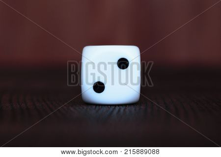Single white plastic dice on brown wooden board background. Six side cube with black dots. Number two.