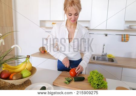 Attractive young woman cuts green cucumber for salad with sharp knife, takes red juicy tomato and several times throws up with smile on face, sniffs freshness of lettuce leaves and takes up shredding vegetables. Blonde at top of modern light kitchen with 
