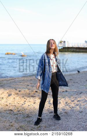 Pretty young woman turns face to camera and looks into camera lens with gentle look and smile, straightens hair with hand and enjoys sea view, standing on shore of blue bottomless sea on clear evening. woman of European appearance with medium brown blond 