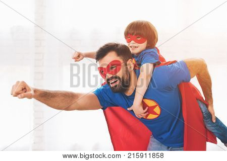 Father and son in the red and blue suits of superheroes. On their faces are masks and they are in raincoats. They are posing in a bright room.