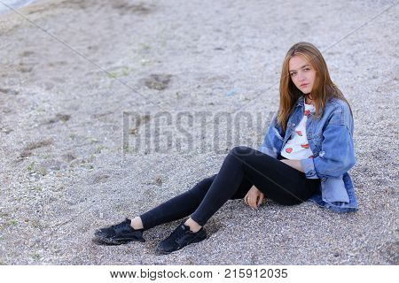 Dreamy girl relaxes and enjoys rest on seaside, throws head back and shakes hair, sitting on sandy beach on Black Sea shore on clear evening. Young European-looking woman with blond mid-length hair dressed in white jacket over which blue denim jacket, bla