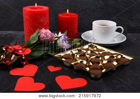 A cup of tea on a stand with a box of chocolates and burning candles, a bouquet of Japanese roses and carved hearts on a stone background