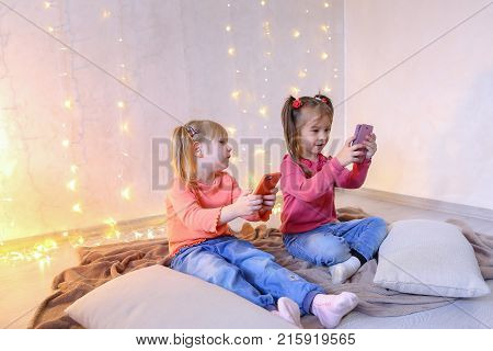 Lovely little girls holding mobile phones and playing games or taking pictures,  typing text for messages on touch screen, they laughing and smiling, sitting on rug in bright room with flickering New Year's garland in evening. Blond girl of European appea