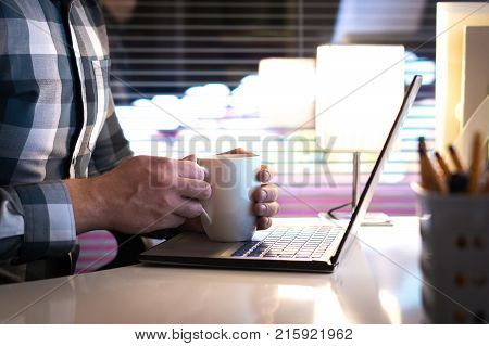 Man holding hands around coffee cup or tea mug in home office late at night. Being sick and drinking hot beverage or having break at work. Laptop on table.