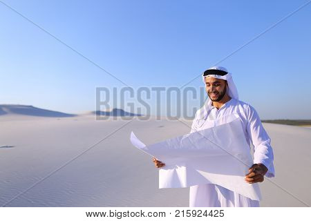 Beautiful Emirate Sheikh businessman studying project of future construction of complex, standing in middle of bottomless desert with white clean sand on clear warm day against blue sky. Swarthy Muslim with short dark hair dressed in kandura, long, spacio
