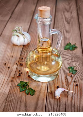 A glass decanter with vegetable oil on a wooden background surrounded by greenery garlic and pepper is closed with a cork
