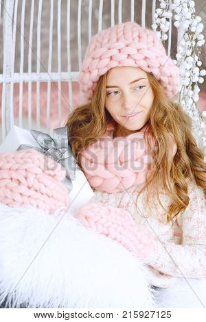 Young woman with red hair and dressed in sweater and knitted pink hat and snood sitting in her favourite chair holding holiday gifts