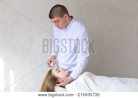 Smart and handsome doctor beautician conducts massage procedure for head of girl who lies on beige couch in medical office. doctor conducts device on hair, assesses condition and gives general recommendations to client, standing in middle of office in cli