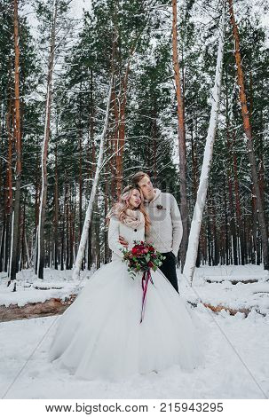 Bride and groom with a rustic bouquet are posing on background of the winter forest. Winter wedding. Artwork