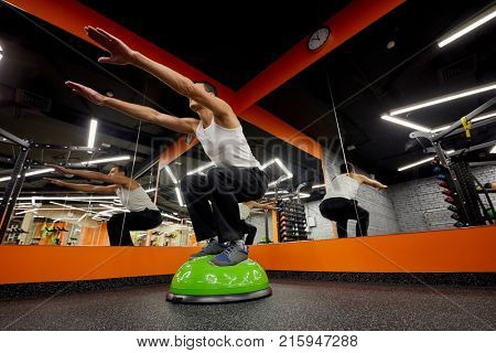 Man doing squatting exercises on inflatable semi ball at fitness studio.