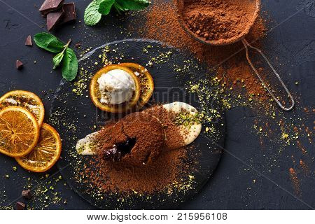 Exquisite french dessert. Chocolate fondant with creme anglaise and vanilla ice cream on round slate with orange citrons. Black background with cocoa, mint and sieve, top view, copy space, top view