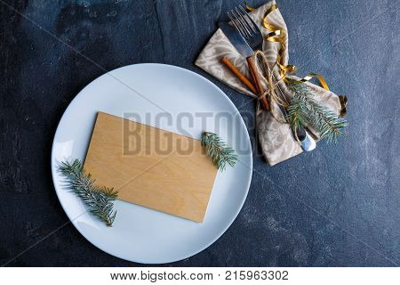 On a white round plate a small plywood board with small fir-trees, next to the cutlery set in a tissue napkin and decorated with fragrant cinnamon sticks, fir-tree and golden ribbon. View from above.