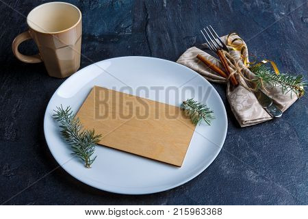 On the table is a white round plate with a small plywood board inside and a fir-tree, next to the serving cutlery are decorated with fir-tree and cinnamon sticks and an empty beige cup.
