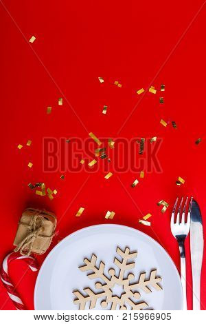 On a red background with a place for an inscription in a white round plate, a decorative wooden snowflake, next to a knife and fork, a small gift box and a Christmas candy. View from above.