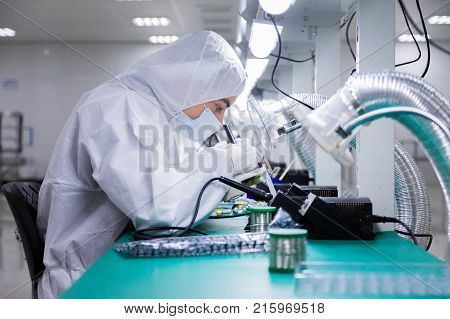 factory workers in white lab suits and face masks, sitting on the chairs are producing tv sets with soldering irons under bright lamps