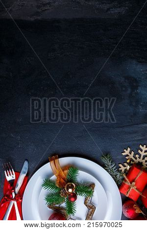 A dark background with a place for the inscription is decorated with a white plate with Christmas decoration and cutlery. View from above.