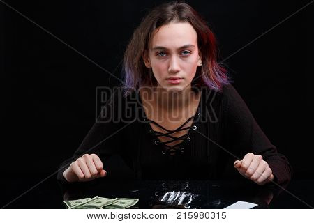 European girl drug addict in a black sweater and with purple hair looks sullen look and sits at the table with a dosed cocaine next to the Dolar bills. On a black background.