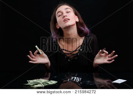 A drug-dependent young girl sniffs cocaine and throws her head back from enjoyment, holding a twisted bank note in her hand. On a black background.