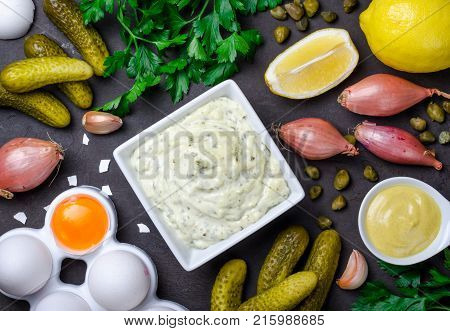 Homemade traditional french sauce remoulade in a white bowl with ingredients lemon eggs shallot onion pickles parsley mustard on a black dark stone slate background. Top view horizontal