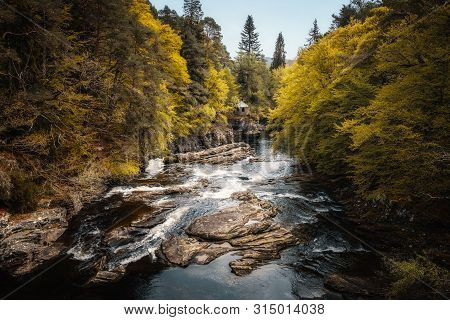 Golden Trees Hang Over The Moriston River At Invermoriston In Scotland With A Small Stone Building I