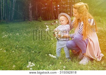 Mom And Her Little Son Love To Relax And Walk In A Summer Park On A Green Flower Meadow. In Their Ha