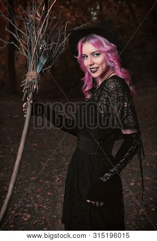 Halloween Theme: Contemporary Smiling Young Witch In Black Dress And Hat With Broom In A Dark Forest