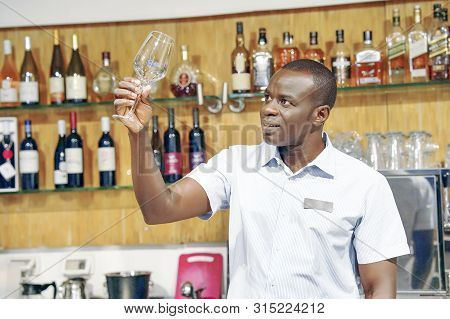 Elderly African Bartender Cleans The Glass. A Handsome Bartender Polishes A Glass Of Wine Glasses. T
