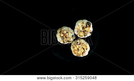 Three Heads Of Garlic With Cut Tops Grilled Rotate On An Isolated Black Background, Top View