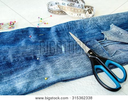 Blue Jeans With Large Hole Folded In Half, Sewing Pins, White Tailor Tape With Centimeters And Inche