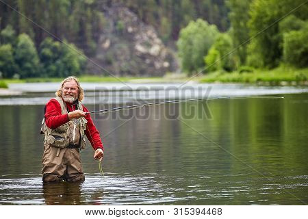 Mature Bearded Whire Wet Man Is Standing In Water In Waterproof Clothing And Preparing To Begin Flyf