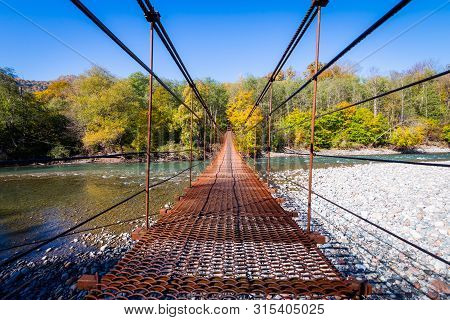 Narrow Metal Foot Bridge Across Mountain River In Autumn.