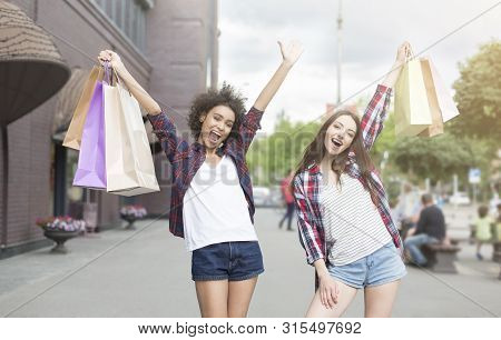 Great Sales In The City. Cheerful Young Ladies Raising Hands Up With Shopping Bags, Walking Down The
