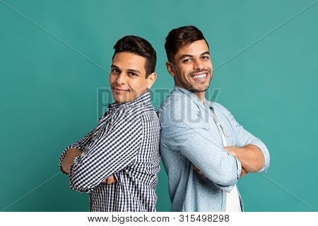 Bromance And Male Bonding. Two Cheerful Hispanic Men Standing Back-to-back On Turquoise Background. 