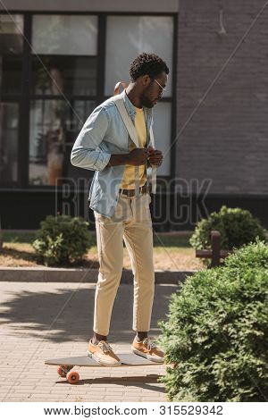 Stylish African American Man Riding Lonboard Along Green Bushes On Sunny Street