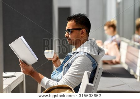 leisure, literature and people concept - indian man reading book and drinking takeaway coffee at street city cafe