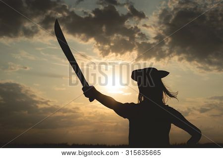 Silhouette Of Pirate Woman In A Pirate Hat And With A Sword In Hands Over A Evening Cloudy Sky Backg
