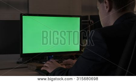 Rear View Of A Young Office Male Worker Sitting At His Desk Working At Computer, Chromakey Monitor. 