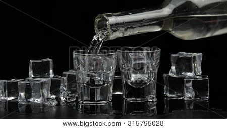 Pouring Up Shots Of Vodka From A Bottle Into Drinking Glass Against Black Background With Ice Cubes.