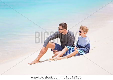 Family Of Two, Father And Son, In Sunglasses And Rashguards Sitting At The Beach Enjoying Beautiful 
