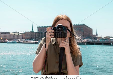 Travel Trip Concept. Turis Blonde Young Girl In A Light Summer Dress Of Green Color Holds A Camera A