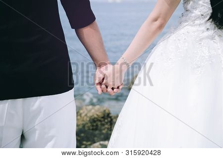 Adorable Wedding Couple Holds Each Other Hands Walking Along The Beach Against The  Ocean Waves In T