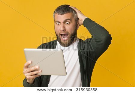 Got Shocking News. Impressed Middle Aged Man Holding Tablet And Touching His Head, Yellow Studio Bac