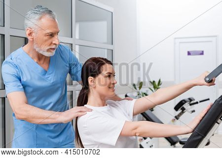 Mature Rehabilitologist Supporting Brunette Woman Working Out In Hospital Gym.