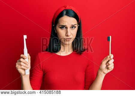 Young hispanic woman choosing electric toothbrush or normal teethbrush skeptic and nervous, frowning upset because of problem. negative person. 